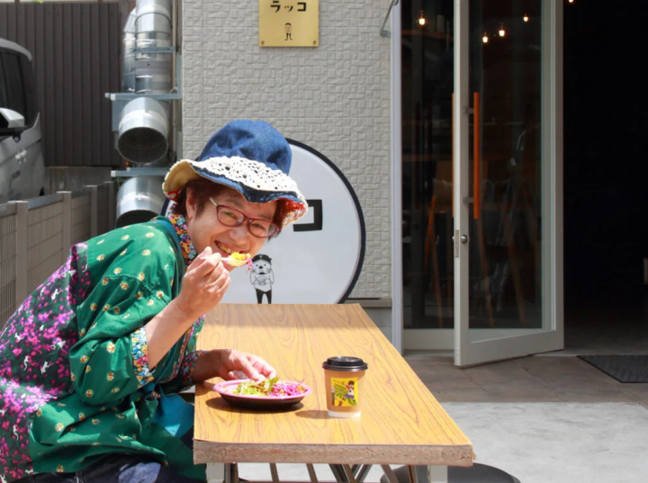 〈ホネカレー〉のカレープレートと〈グリーンハンズコーヒー〉のホットコーヒーを楽しむマダム。カメラを向けるといい笑顔